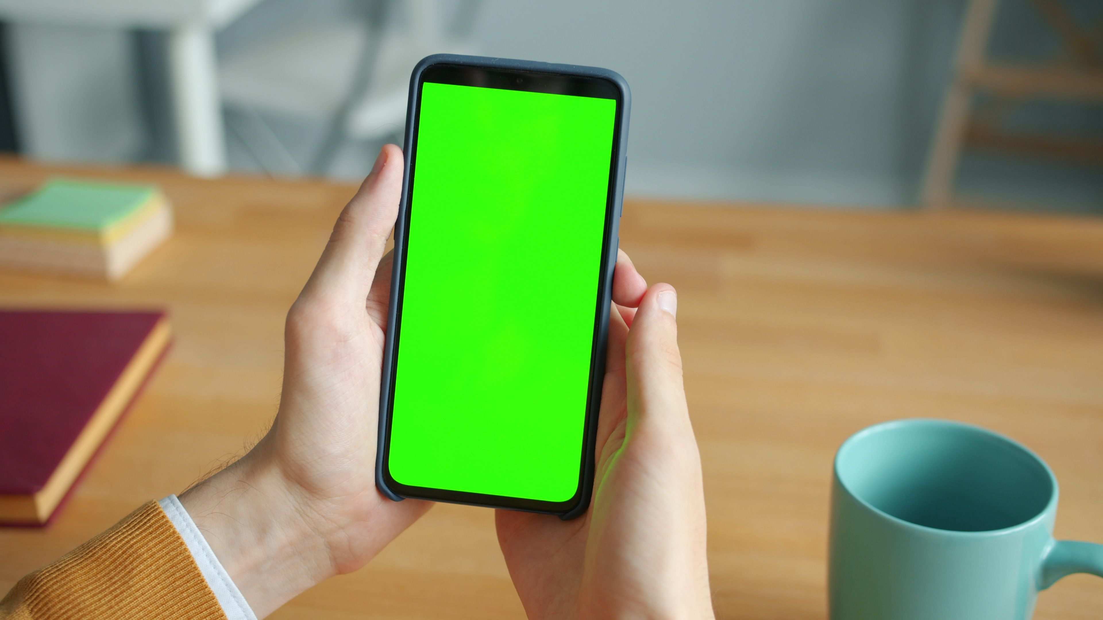 มือถือแสดงหน้าจอแอปพลิเคชันขณะทดสอบ — Close-up of a person holding a smartphone with a green screen and a mug on a wooden table.