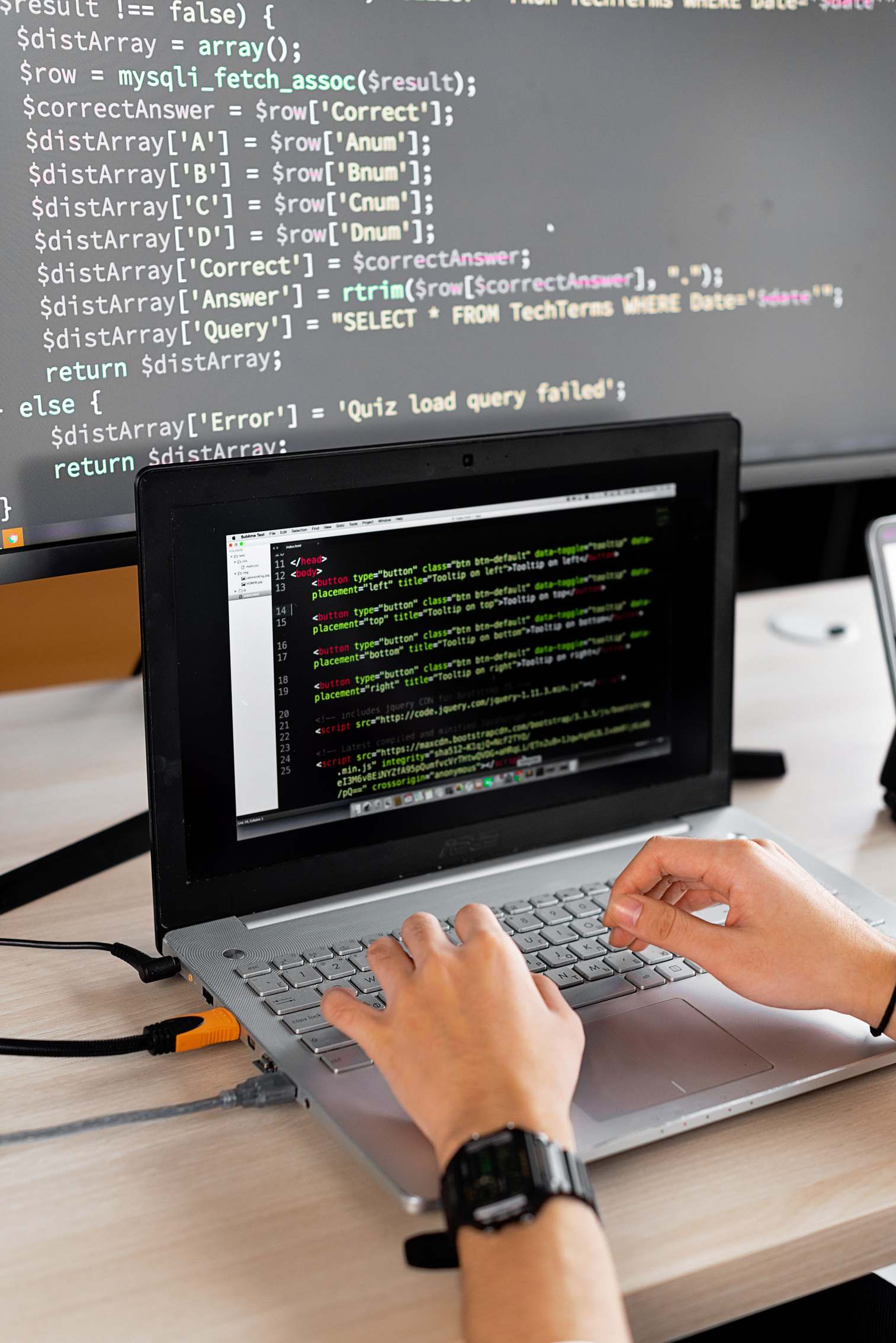 หน้าจอแสดงโค้ดระหว่างตรวจทานงาน — Person coding at a desk with laptop and external monitor showing programming code.